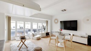 A modern living room with a large TV, a dining area with a white table and chairs, and a cozy seating area with a sofa and a coffee table. - Cozy Studio - 2P - Place de la Bourse (Paris)