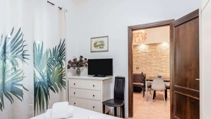 A hotel room with a bed, a TV, a desk, and a chair. There is a door leading to a dining area.