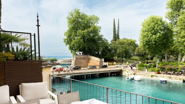 A terrace with a table set for dining, overlooking a canal with boats and a bridge. - Cordigero - Riverside Suite (Peschiera del Garda)