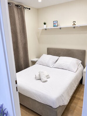 A neatly made bed with white linens and two pillows, a bedside table with a lamp, a shelf with a plant and picture, and a brown curtain.