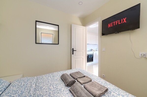 A hotel room with a bed, a wall-mounted TV displaying 'NETFLIX', and a door leading to a kitchenette.