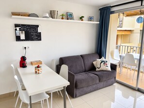 A modern living room with a grey sofa, white chairs, a small table with a red kettle, a chalkboard with writings, and a balcony with a blue curtain.