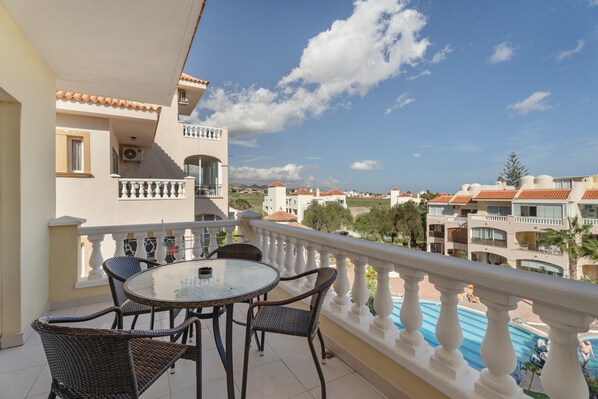 A balcony with a round table and wicker chairs, overlooking a swimming pool and other buildings.
