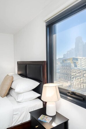 A hotel room with a bed, a nightstand, a lamp, and a window with a city view. - Heritage Top Floor Loft CBD Parking (Melbourne)