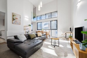 A modern living room with a black leather sofa, a dining area with a wooden table, and a large window providing natural light.