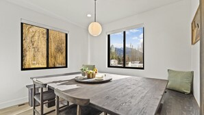 A spacious dining area with a large wooden table, two windows with mountain views, and a hanging light fixture. - Glacier Retreat Estates - Sweeping Mountain Views! (Columbia Falls)