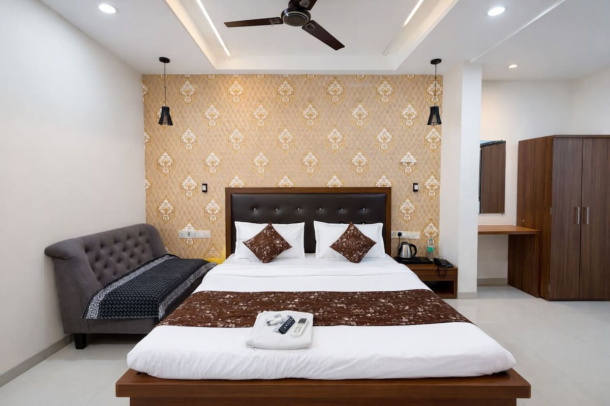 A modern hotel room with a large bed, a grey armchair, a wooden wardrobe, and a ceiling fan.