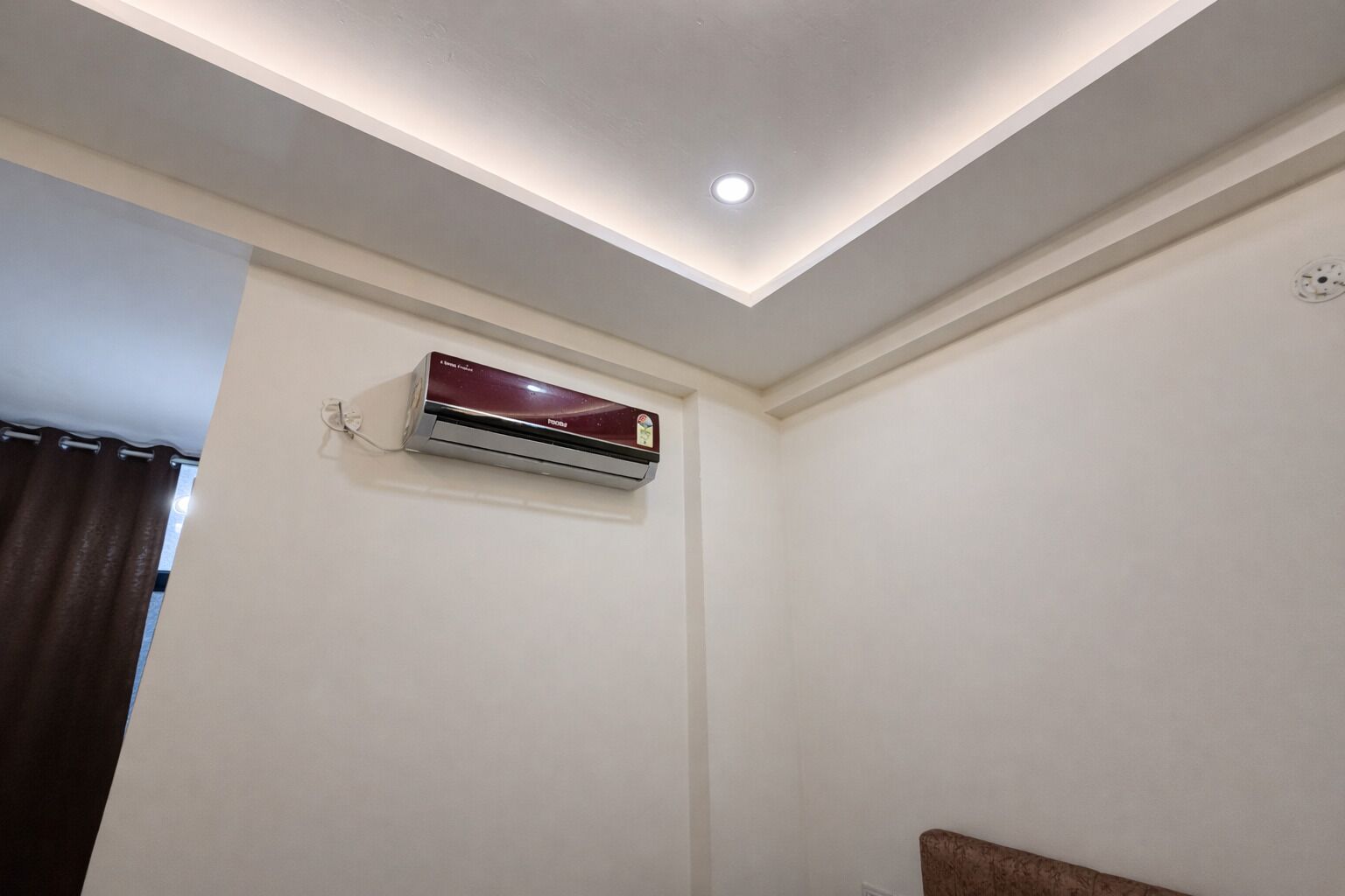 A room with a ceiling light, an air conditioning unit mounted on the wall, and a window with curtains.