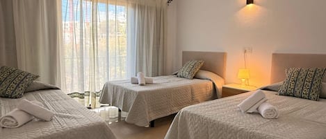 A hotel room with three beds, each with a white quilted bedspread and matching pillows. There is a window with sheer curtains, a bedside lamp, and an air conditioning unit mounted on the wall.