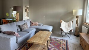 Una sala de estar con un sofá seccional gris, una mesa de centro de madera, un tapete estampado, un sillón blanco de pelo y una lámpara alta.