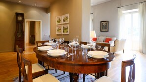 Une salle à manger avec une table en bois dressée pour six personnes, entourée de chaises. On aperçoit un canapé et une lampe au fond de la pièce.
