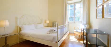 A bedroom with a white metal bed, a bedside table with a lamp, a radiator, a window with curtains, and a glass cabinet.
