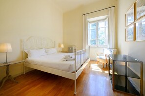 Une chambre avec un lit en métal blanc, une table de chevet avec une lampe, un radiateur, une fenêtre avec des rideaux et une vitrine.