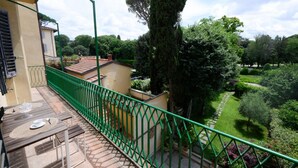 Un balcon doté d’une balustrade en métal vert, une table avec un set de table, et une vue sur un jardin avec des arbres et un sentier.