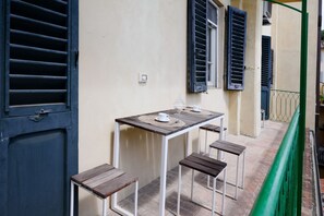 Un petit espace extérieur aménagé pour prendre les repas, avec une table et quatre tabourets en bois, une rambarde verte et un bâtiment doté de volets.