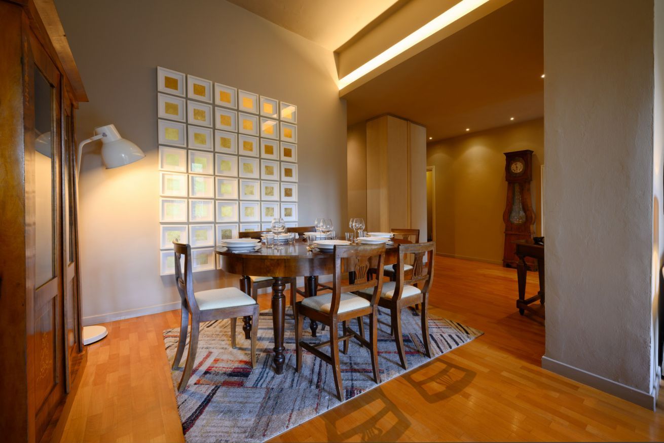 A dining area with a wooden table set for six, surrounded by chairs. A patterned rug is on the floor, and a wall-mounted light fixture is above the table.