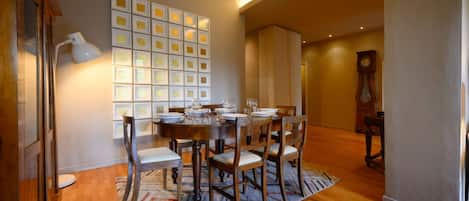 A dining area with a wooden table set for six, surrounded by chairs. A patterned rug is on the floor, and a wall-mounted light fixture is above the table.