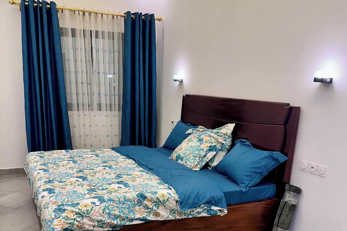 A neatly made bed with a floral bedspread, blue pillows, and a wooden headboard in a room with tiled flooring and a window with blue curtains.