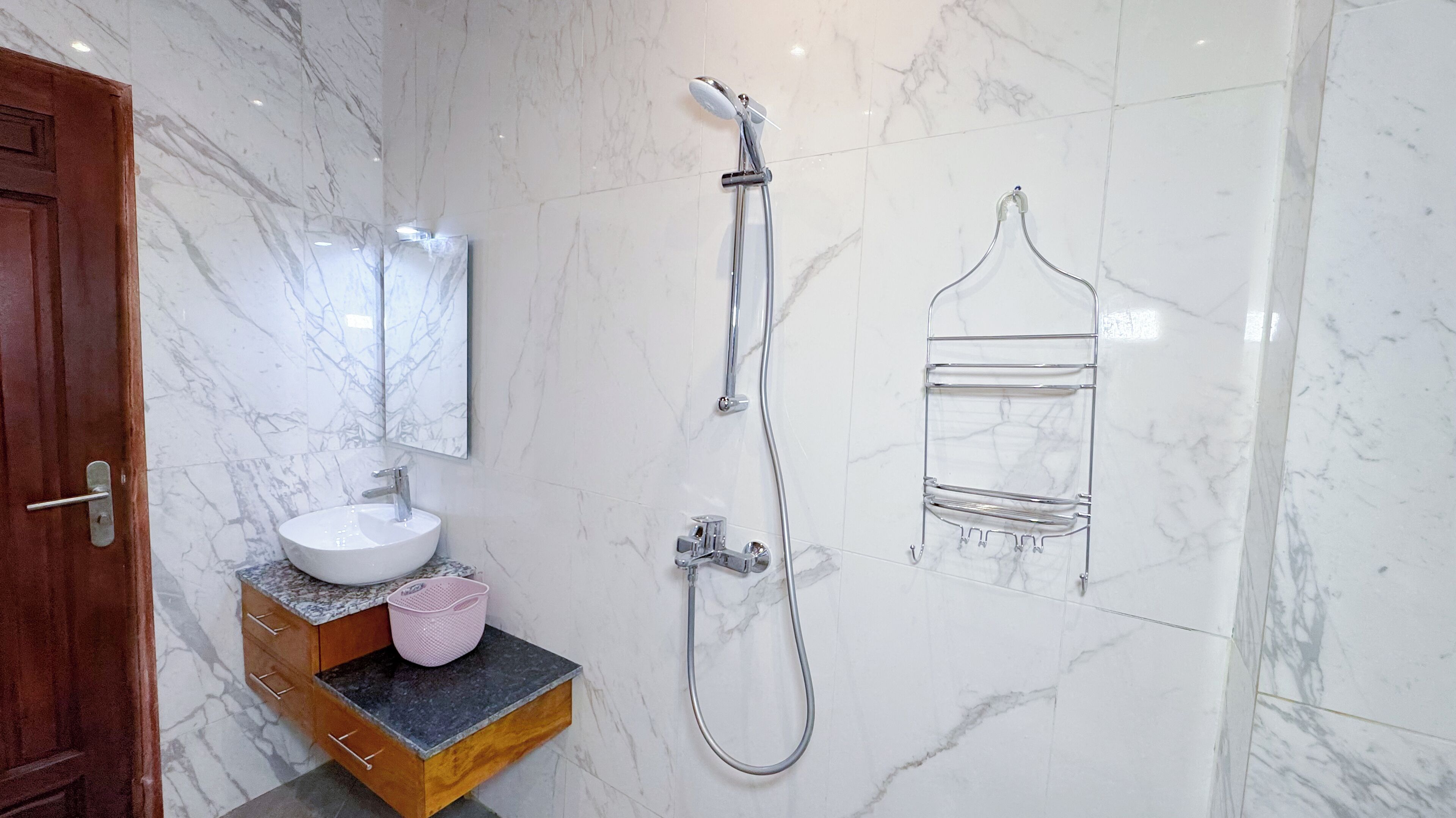 A bathroom with a marble wall, a wooden vanity, a white sink, a mirror, a shower with a handheld showerhead, and a towel rack.