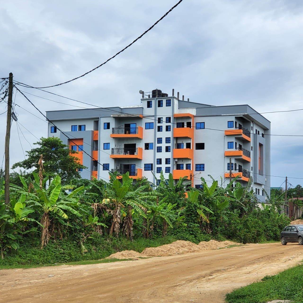 Un immeuble à plusieurs étages doté de balcons, entouré d’une végétation luxuriante et bordé d’un chemin de terre.