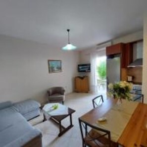 A compact living space with a sofa, a dining table, and chairs. A television is mounted on the wall, and there is a window providing natural light. - Prokimea Seaview Apartments (Lefkada)