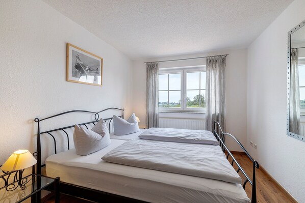A double bed with a headboard, a bedside table with a lamp, a window with curtains, and a framed picture on the wall.