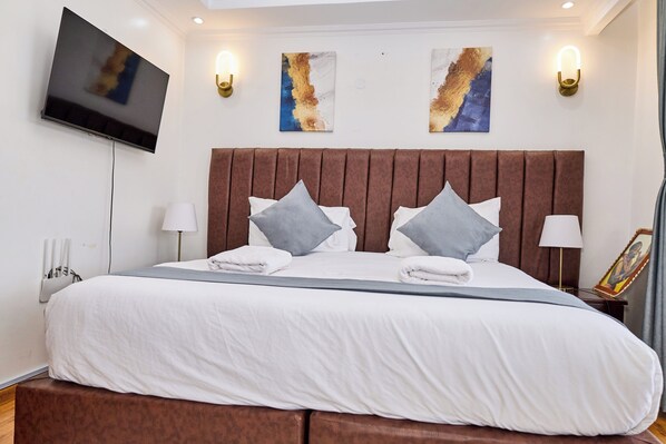 A neatly made bed with pillows, a flat-screen TV mounted on the wall, and two wall-mounted light fixtures.