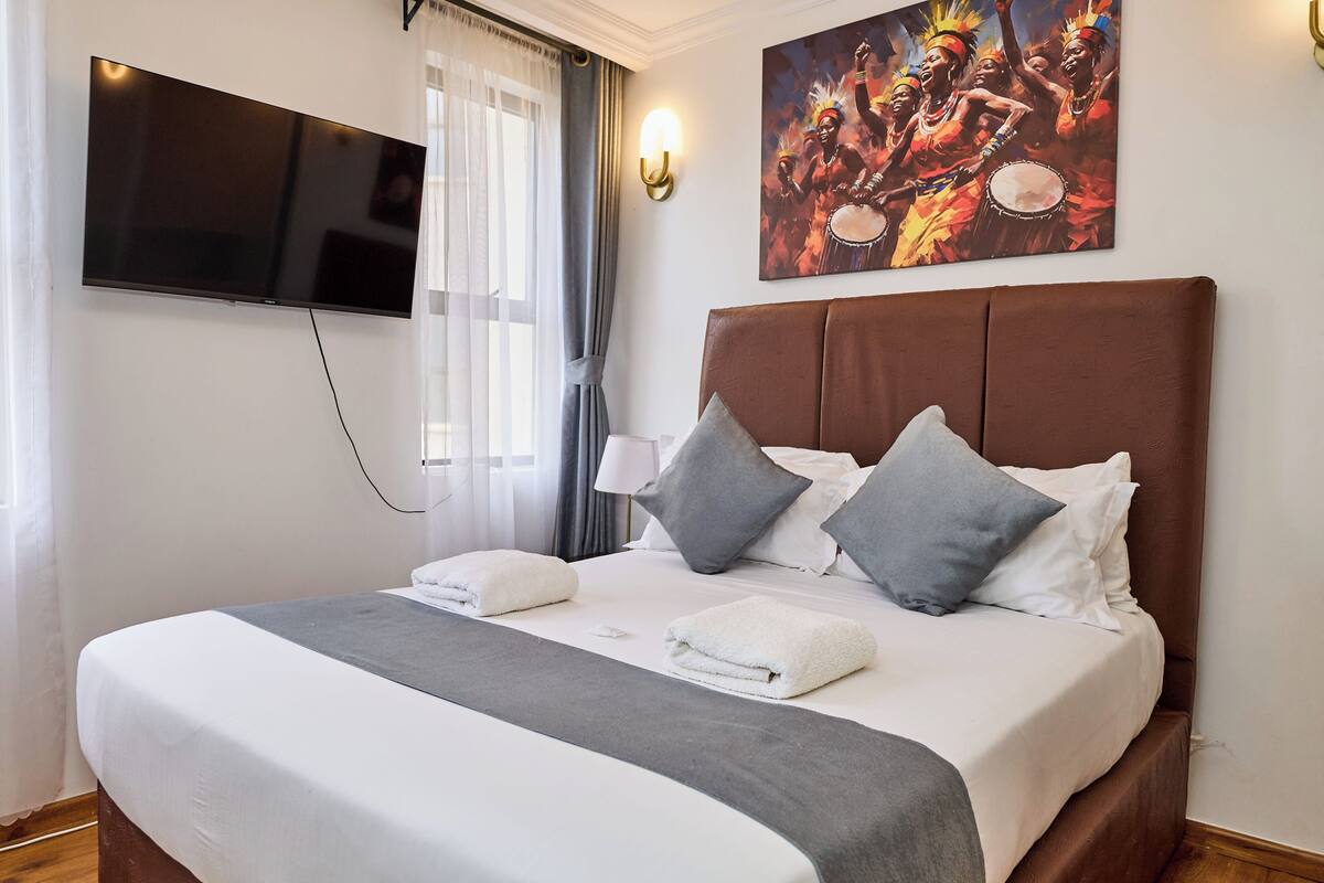A neatly made bed with pillows, a flat-screen TV mounted on the wall, and a painting above the bed.