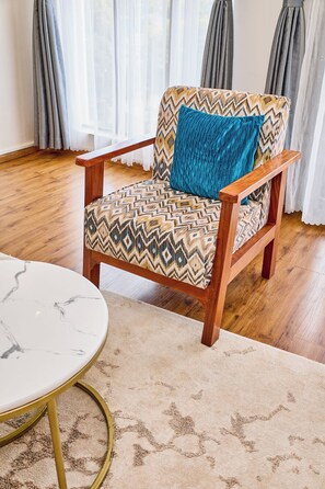 A wooden armchair with a patterned fabric and a blue pillow, a round table with a marble top, and a beige carpet with a subtle pattern. - Sangara by Edmor Suites. (Nairobi)