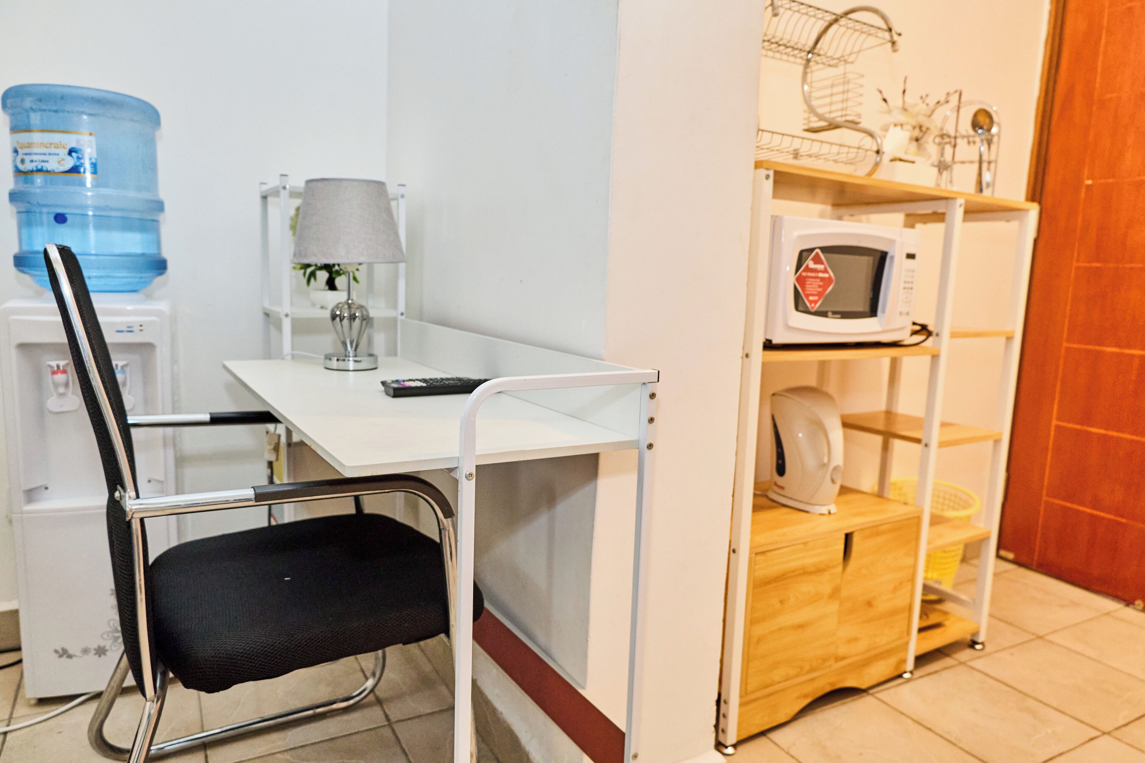 A compact kitchenette with a white countertop, a black chair, a water dispenser, a microwave, and a small wooden cabinet.