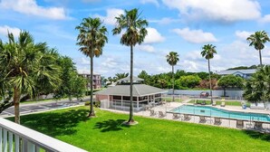 Uma área de piscina com um deck cercado, palmeiras e um edifício com telhado cinza.