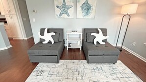Uma sala de estar moderna com dois sofás cinza, um tapete branco e duas almofadas em formato de estrela-do-mar. Há duas obras de arte penduradas na parede retratando uma estrela-do-mar e uma concha. Uma luminária de chão e uma mesinha lateral branca também estão visíveis.
