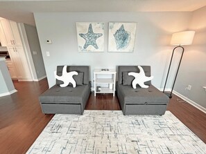 Una sala de estar moderna con dos sofás grises, un tapete blanco y dos cojines con forma de estrella de mar. Se observan dos obras de arte colgadas en la pared que representan una estrella de mar y una concha marina. También se aprecia una lámpara de pie y una mesita auxiliar blanca.