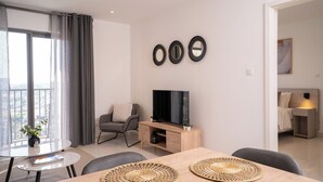 A modern living room with a flat-screen TV, a dining table with woven placemats, a grey sofa, and a large window with curtains. - One bedroom Luxury @ Loxwood…..Enjoy! (Accra)