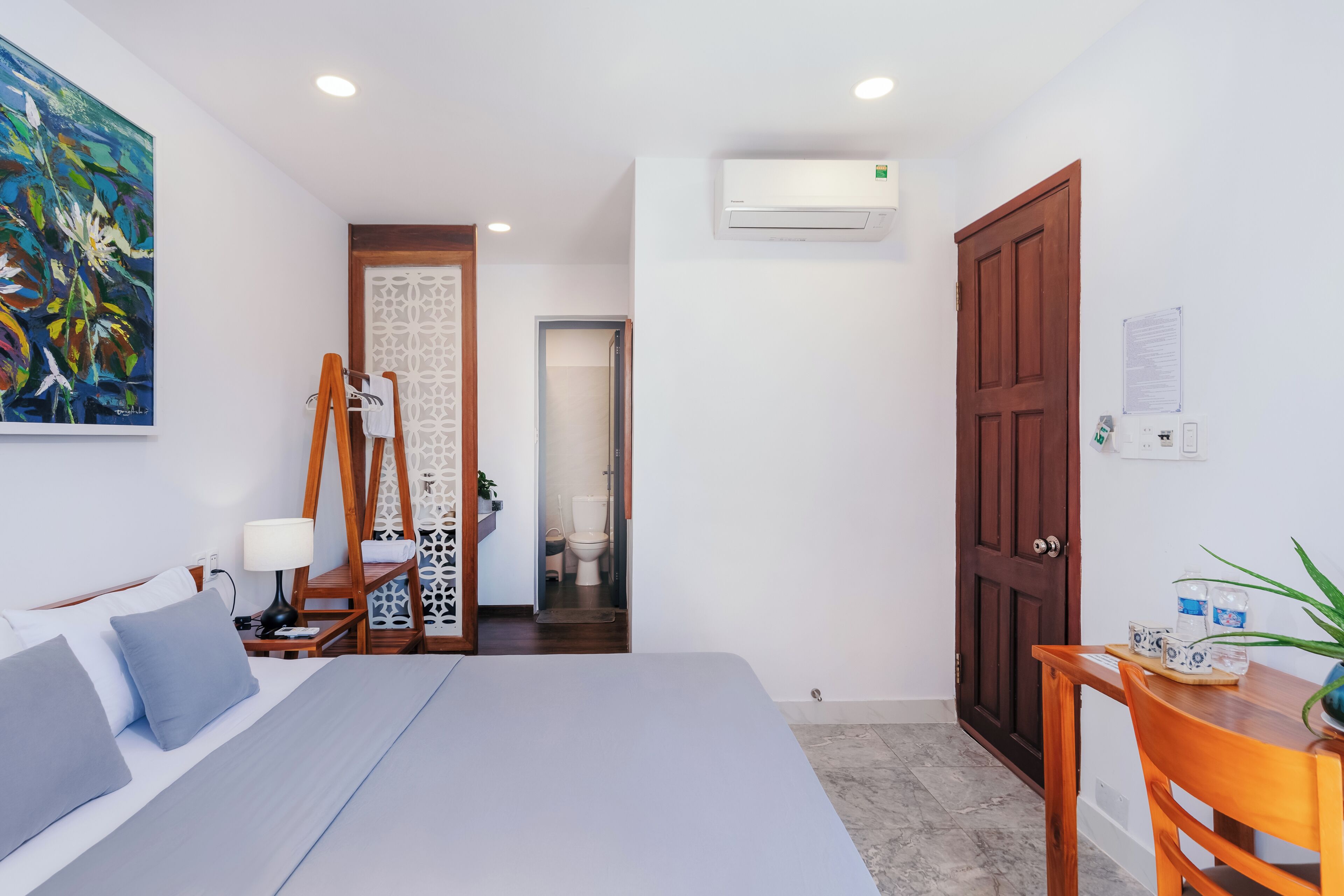 A hotel room with a bed, a desk, a chair, an air conditioner, a wooden door, and a painting on the wall.