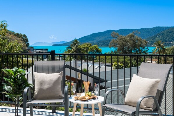A balcony with two chairs, a small table with a bottle, glasses, and snacks, overlooking a turquoise sea and green hills.