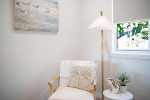Una habitación con una silla blanca, una lámpara de pie, un cuadro, un ventanal con vistas y una pequeña mesa con una planta y adornos.