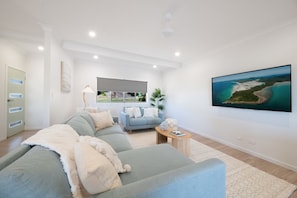 A modern living room with a sectional sofa, a small side table, a flat-screen TV mounted on the wall, and a large window providing a view of the outdoors. - Orana Coastal Retreat (Airlie Beach)