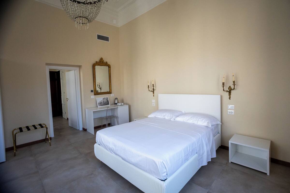 Un dormitorio con una cama grande, un escritorio, una silla, un banco, un espejo y apliques de pared.