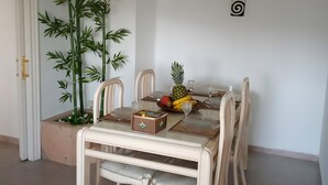 Un coin repas avec une table dressée pour quatre personnes, des chaises, une plante en pot et un panier de fruits.