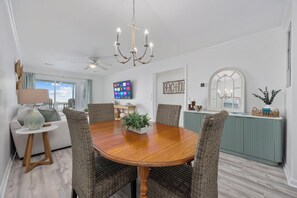 Un coin repas avec une table en bois, des chaises en osier, un ventilateur de plafond, un lustre et un téléviseur fixé au mur.