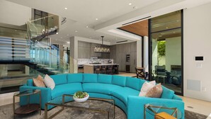 A modern living room with a teal sectional sofa, a glass coffee table, and a staircase with glass railings. - Luxury Coastal Escape w/ Resort-Style Amenities & Stunning Views (Del Mar)