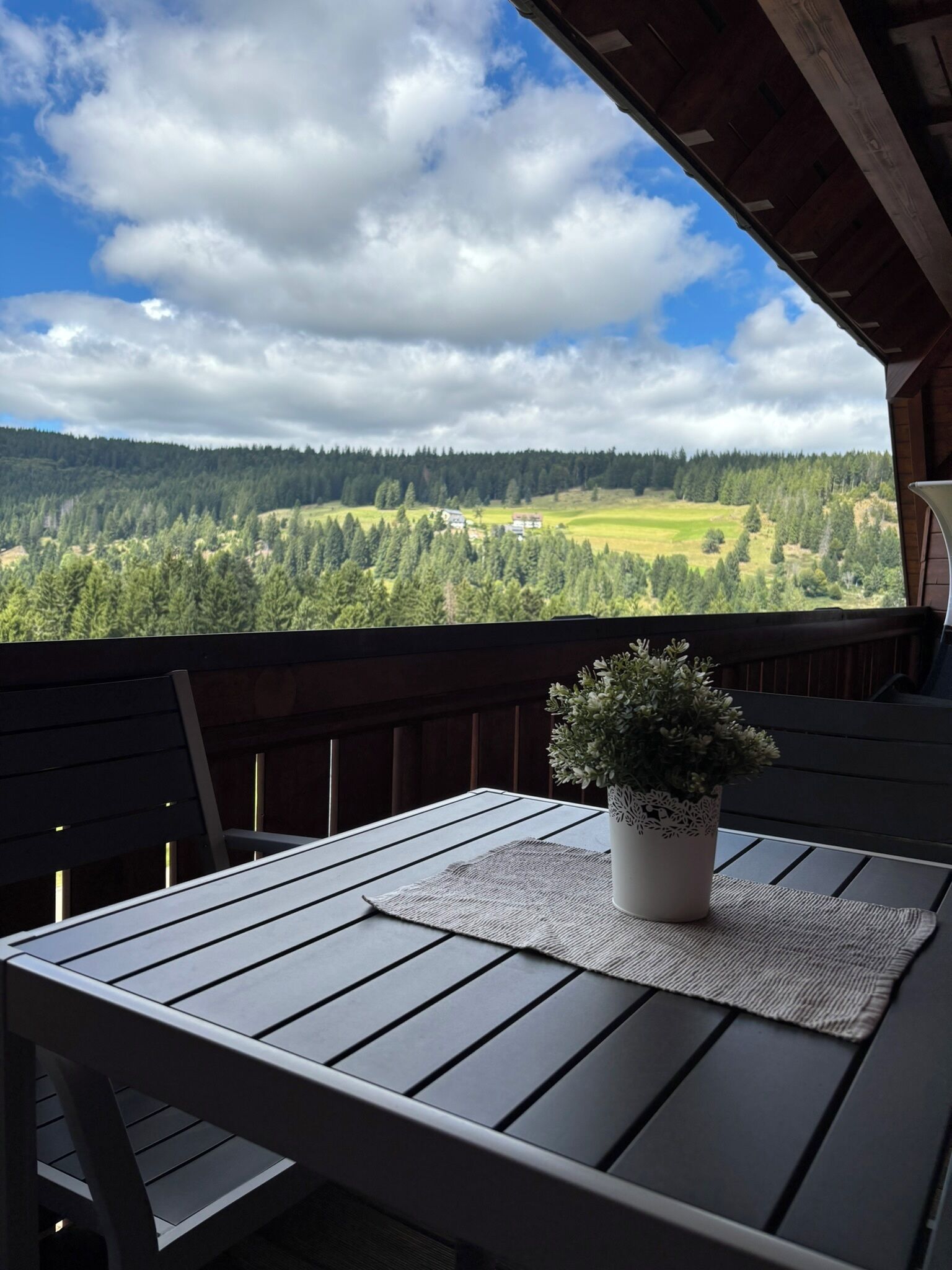 A wooden deck with a table and chairs, overlooking a forested valley with a clear sky.