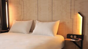 A neatly made bed with white linens and pillows, a bedside table with a lamp, and a wall with a geometric wood panel design. - BB HOTEL Carcassonne Rocadest La Cite (Carcassonne)