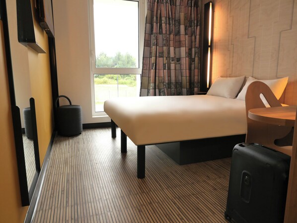 A modern hotel room with a large bed, a desk with a chair, a suitcase, and a view of greenery outside. - BB HOTEL Carcassonne Rocadest La Cite (Carcassonne)