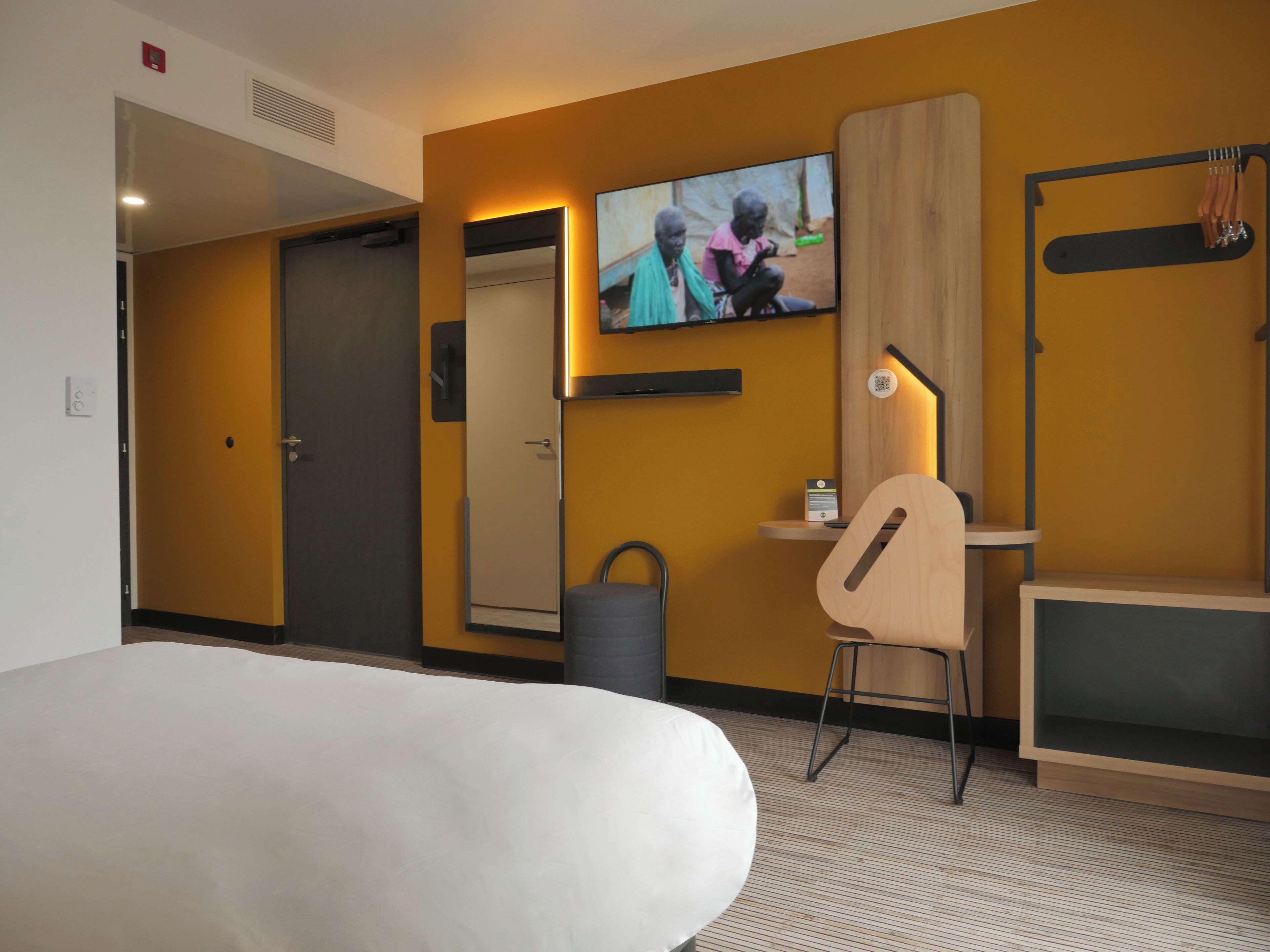 Una habitación de hotel con cama, un escritorio con silla, un televisor fijado a la pared y un armario de madera.