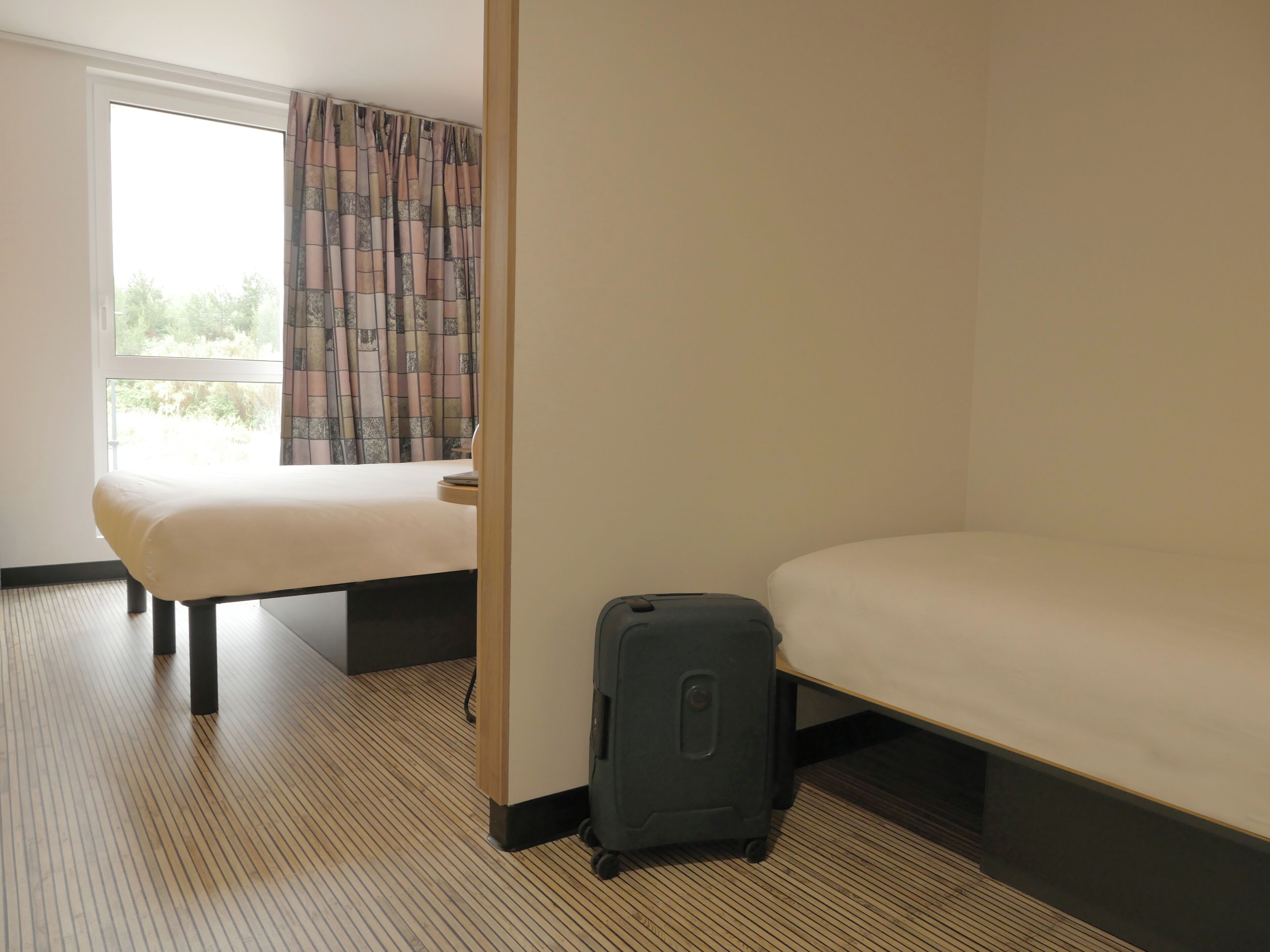 Una habitación de hotel con cama, maleta y ventana con cortinas.