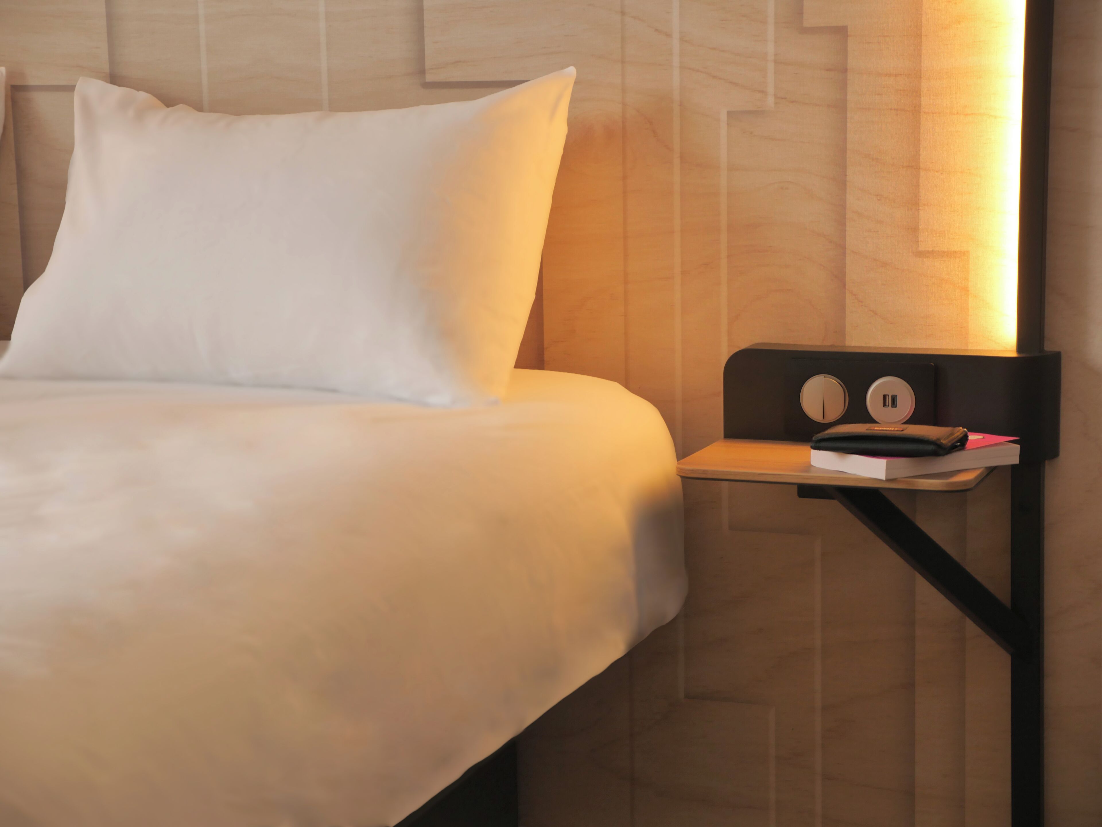 A neatly made bed with white linens and a single white pillow. A small bedside table with a black frame holds a book, a wallet, and a pair of earphones. The wall behind the bed features a wooden panel design.