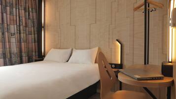 Une chambre d’hôtel comprenant un lit, un bureau avec un ordinateur portable, une chaise, un porte-manteau et un mur orné d’un motif géométrique.