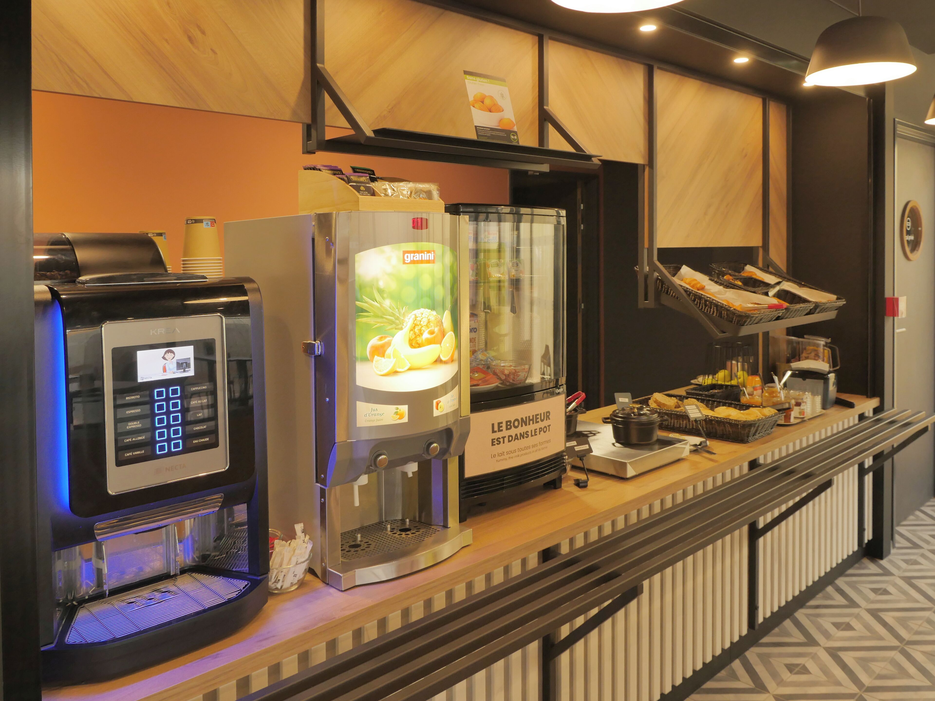 A self-service coffee station with a digital coffee maker, a granini coffee dispenser, and a variety of food items on a counter.